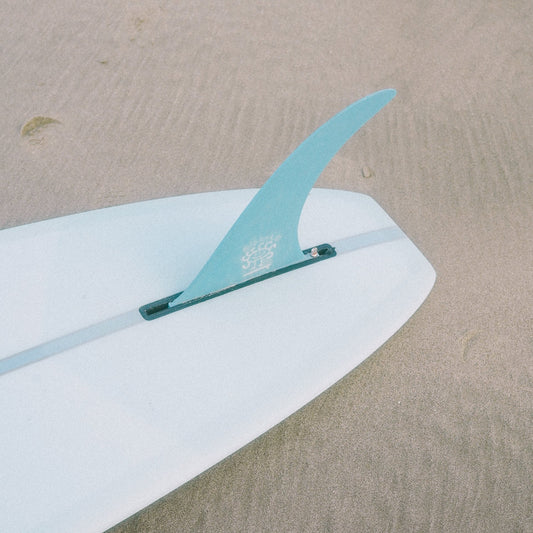 A surfboard with a vibrant blue fin is resting on the sandy beach. The surfboard is positioned diagonally, with the blue fin prominently visible. The background features a stretch of golden sand. The associate product is a digital gift card of an amount of your choosing.