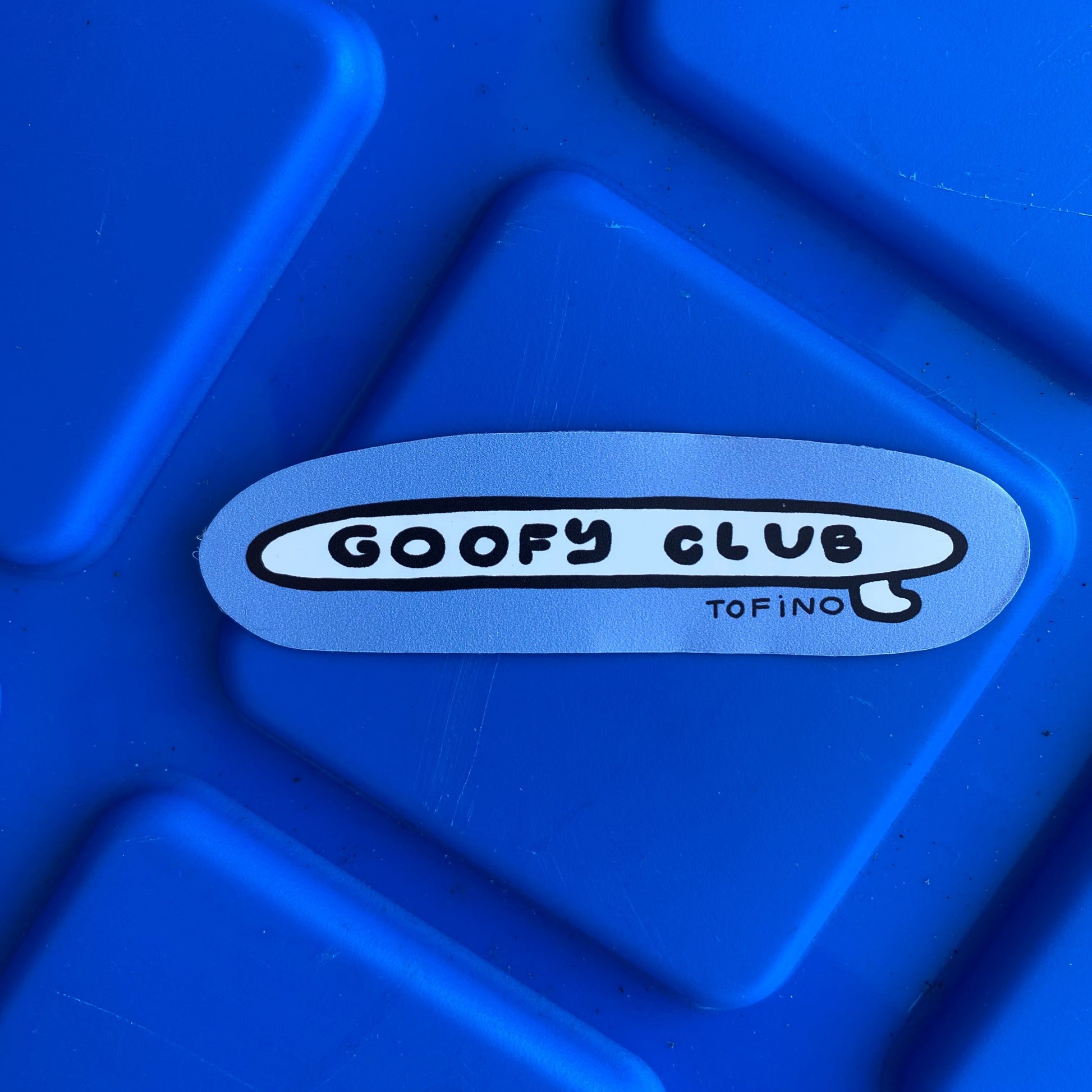 A close up of a sticker on a blue surface. The sticker reads "GOOFY CLUB" in black text on a white background in a surfboard design. 