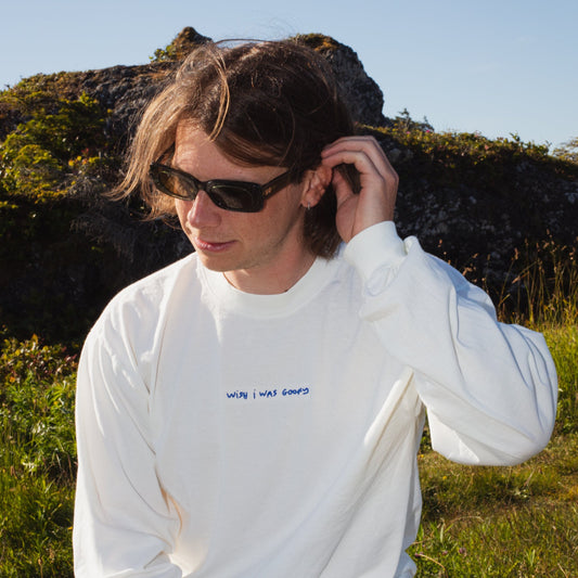 A person with medium length brown hair wearing sunglasses wears a while long sleeve with small blue embroidered text that reads"wish I was goofy". 