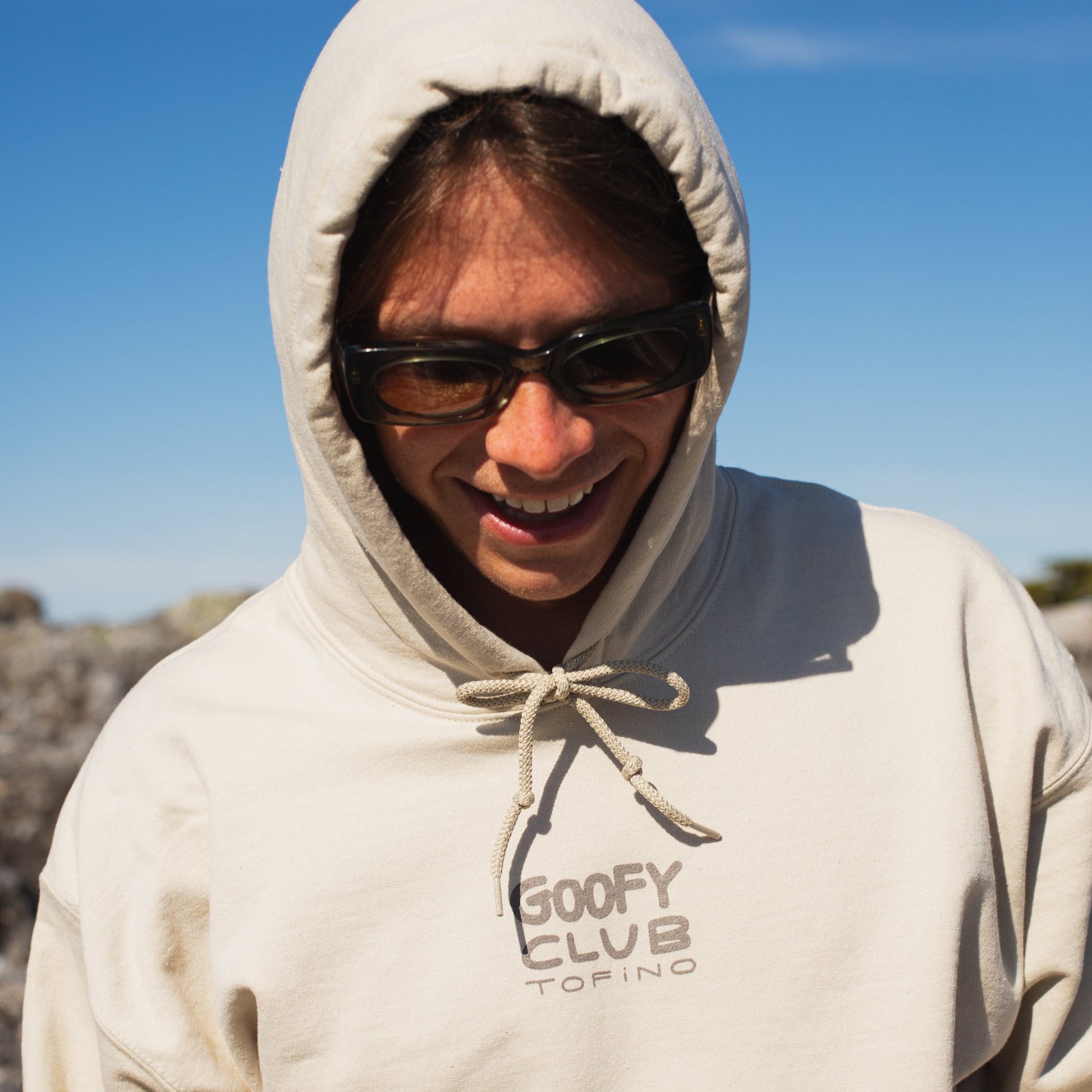 A smiling person wears an off-white coloured hoodie with the hood up. The words "GOOFY CLUB TOFINO" illustrate the front of the hoodie. 