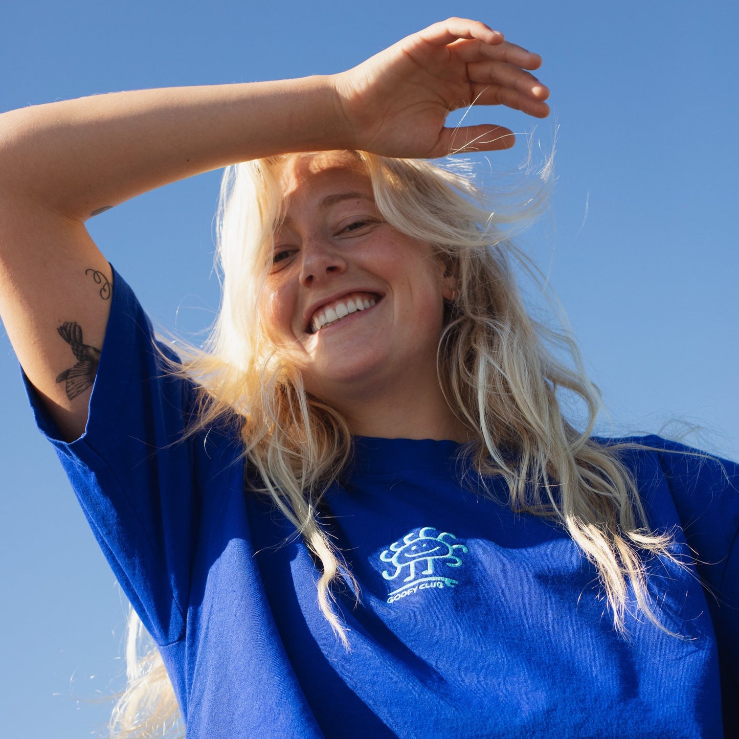 A smiling person with their hand shielding there eyes from the sun wears a blue  t-shirt with a small smiling sun character embroidered on the front of the tee in white thread. 