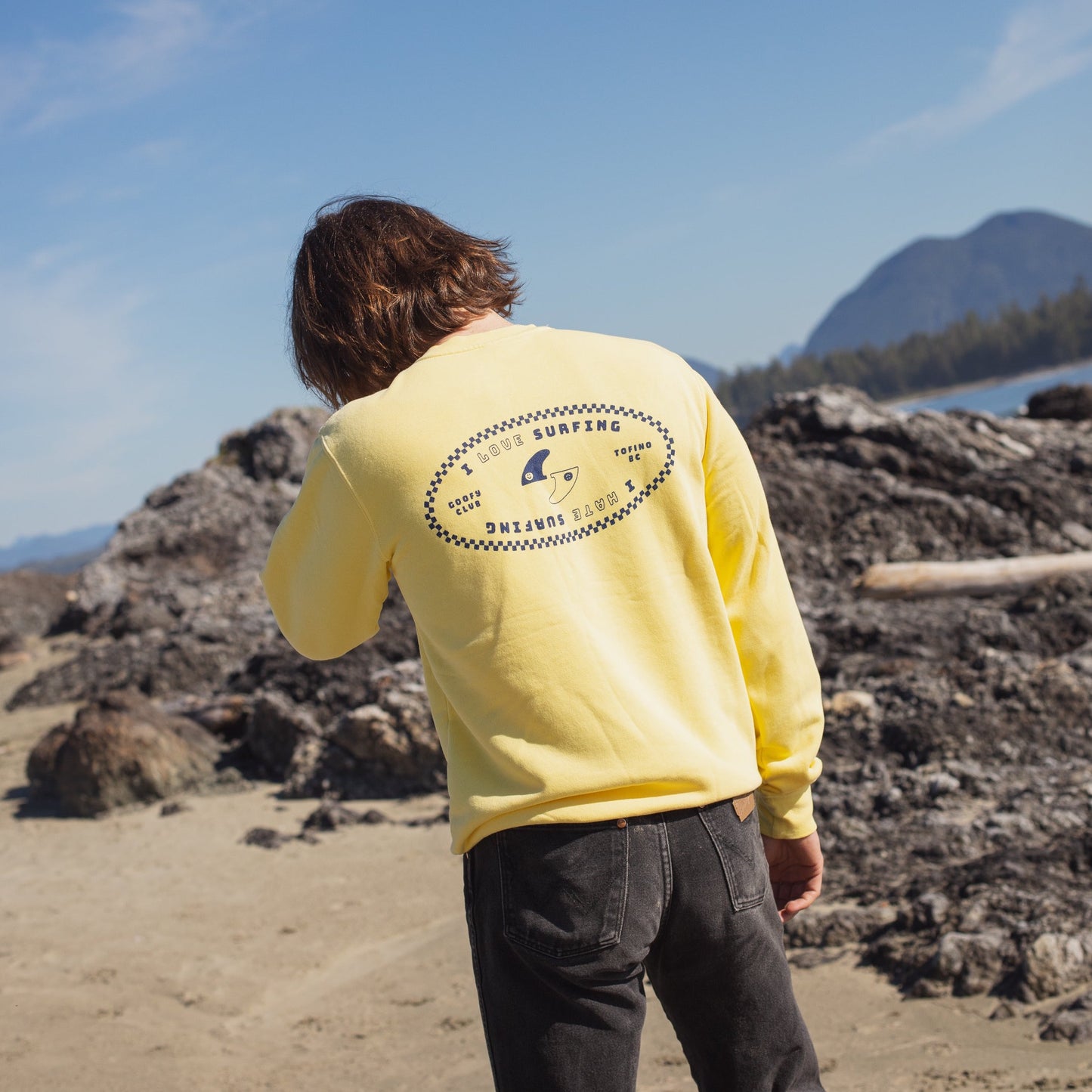 A person with shoulder-length brown hair, seen from behind, standing on a rocky beach with a backdrop of mountains and blue sky. They are wearing a yellow long-sleeve shirt with a circular blue graphic on the back that features surfing-related imagery and text that reads 'SURFING ORIGINAL CLUB.