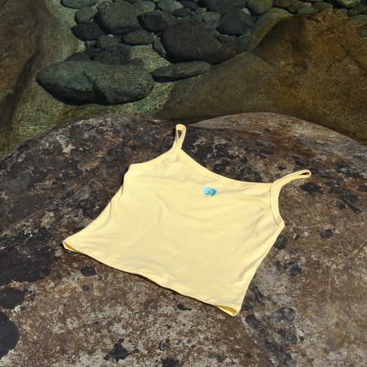 A yellow tank top lays on a rock near a pool of water. The butter-yellow tank features an embroidered blue spiral embroidered on the front.