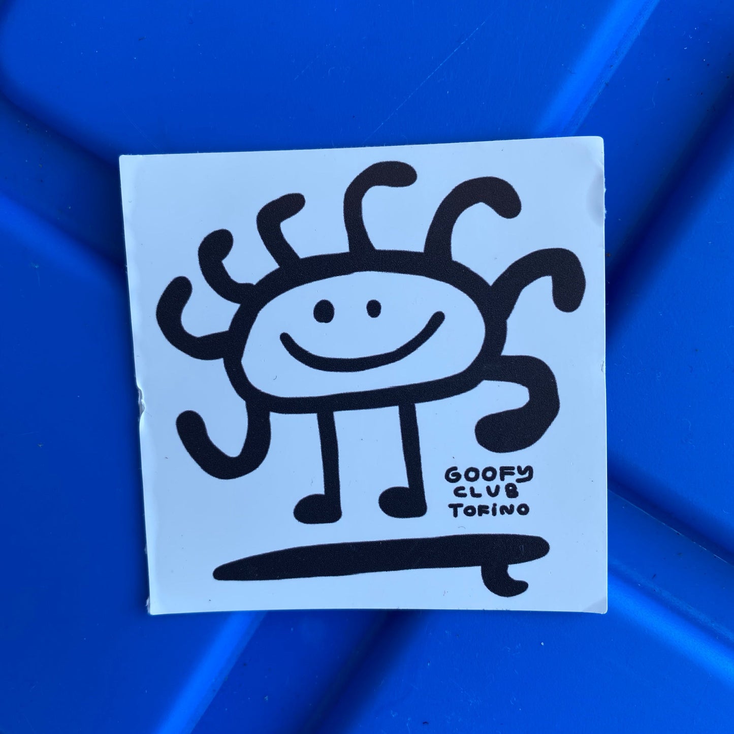 A small square light blue sticker with a smiling sun character and "Goofy Club Tofino, BC