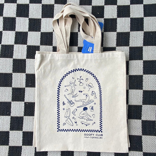 Canvas tote bag laid on a black and white checkered surface. The bag features a blue graphic on the front, depicting a variety of small, simple illustrations of surfers and surfing-related objects within an arched frame. Below the graphic, the text reads 'GOOFY CLUB - TOFINO, BC