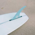 A surfboard with a vibrant blue fin is resting on the sandy beach. The surfboard is positioned diagonally, with the blue fin prominently visible. The background features a stretch of golden sand. The associate product is a digital gift card of an amount of your choosing. 