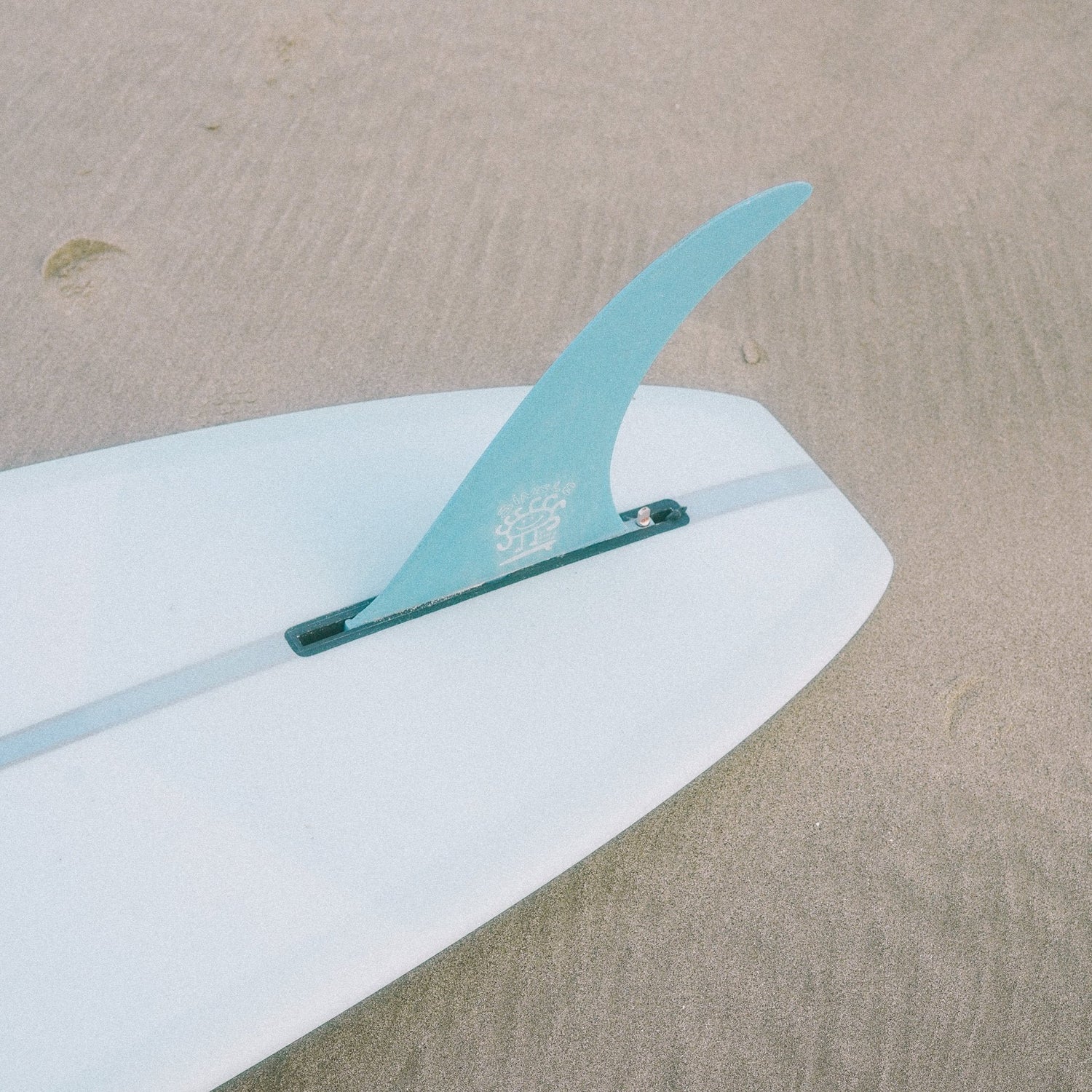 A surfboard with a vibrant blue fin is resting on the sandy beach. The surfboard is positioned diagonally, with the blue fin prominently visible. The background features a stretch of golden sand. The associate product is a digital gift card of an amount of your choosing. 