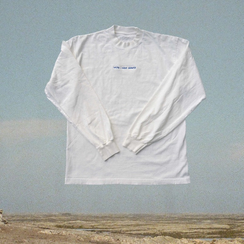 A plain white long sleeve shirt laid flat on a neutral beach background. The shirt has small blue embroidered text centred on the chest that reads "wish I was goofy". 