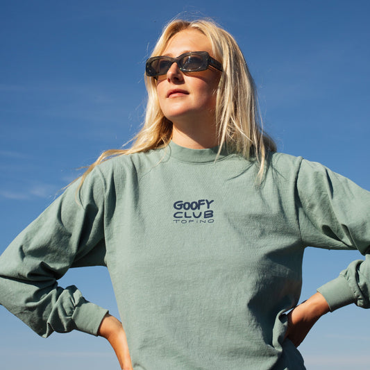 A person standing with their hands on their hips wears sunglasses and a seaform, sage green coloured long sleeve shirt. The words "GOOFY CLUB TOFINO" illustrate the front of the shirt.