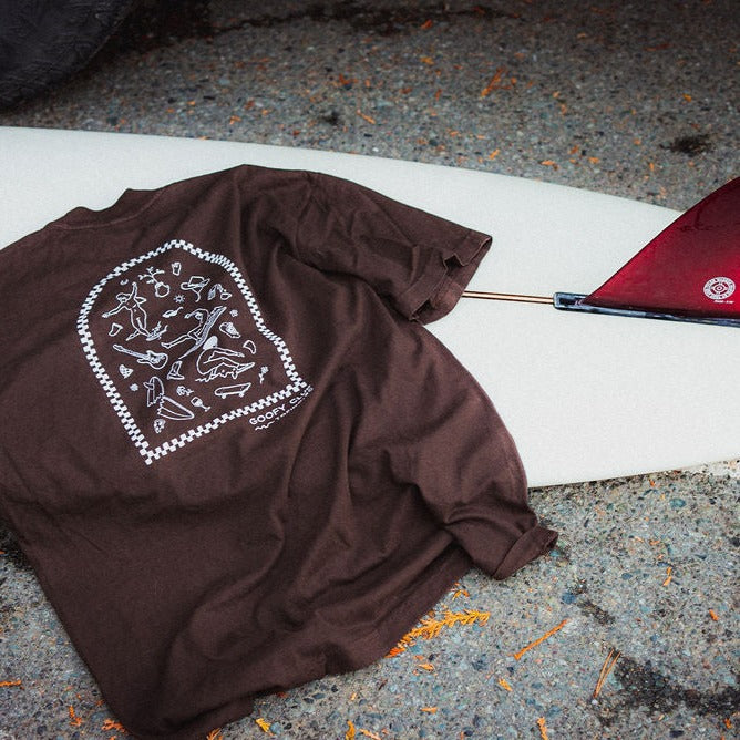 A warm brown t-shirt lays flat across a surfboard. The tee feature a white graphic on the back, depicting a variety of small, simple illustrations of surfers and surfing-related objects within an arched frame. Below the graphic, the text reads 'GOOFY CLUB - TOFINO, BC.'