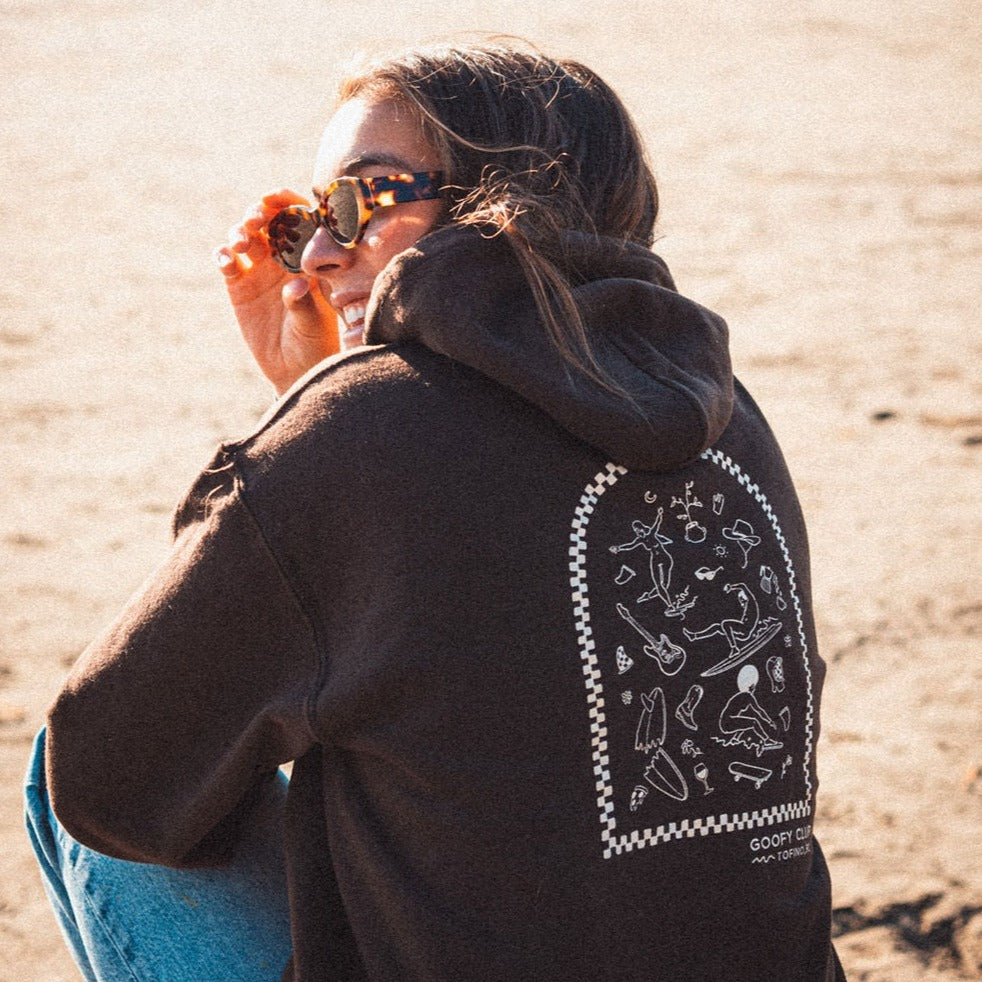 A person with their back to the camera sits on the beach. They are wearing a brown hoodie with a white graphic on the front, depicting a variety of small, simple illustrations of surfers and surfing-related objects within an arched frame. Below the graphic, the text reads 'GOOFY CLUB - TOFINO, BC.'